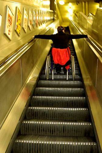 Woman using a wheelchair riding on an escalator to the upper level of a building.
