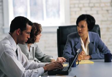 three people talking at a table, one man using a laptop computer