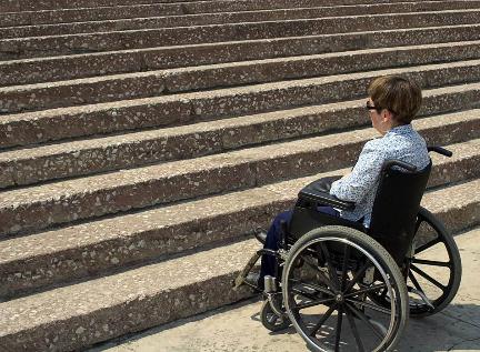 A woman in wheelchair at bottom of stairs looks at an inaccessible building.
